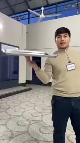 Here's a description of the video:
A man is standing in a large, open room with a tiled floor and a ceiling fan. He is holding a rectangular metal tray in his hand and balancing it on his fingertip. He rotates the tray, showing it from different angles. The room appears to be part of a building with windows and doors. The sky is visible through the roof, indicating it might be outdoors or a covered area. The man wears a light-colored, zip-up sweater, dark jeans, and a badge labeled "Volunteer." There is a walkie-talkie clipped to his pants. A fire extinguisher is mounted on the wall behind him.