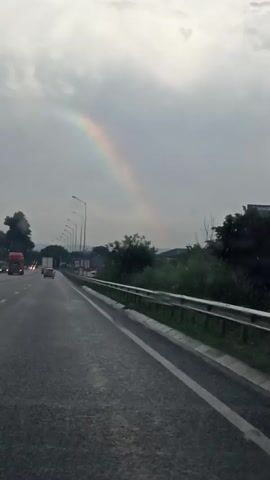 The video shows a view from inside a vehicle traveling on a wet highway under a cloudy sky. A faint rainbow is visible in the distance, arcing across the sky. The highway has multiple lanes, with other vehicles visible in the distance. On the right side of the highway, there is a metal guardrail, followed by greenery and some buildings. The road surface appears wet, suggesting recent or ongoing rain. The lighting is dim, consistent with an overcast day.