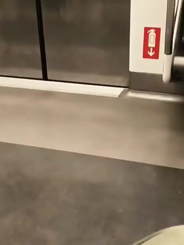 The video captures the interior of a metro train, identified as being in Mumbai, India. The scene opens with a close-up of a metallic train door, on which a red fire extinguisher symbol is prominently displayed. A passenger in a striped shirt and dark pants is seen sitting nearby, preoccupied with their mobile phone. Their feet are clad in pink flip-flops, and a blue bag rests on the floor next to them.

As the camera pans to the right, a wider view of the metro carriage is revealed. The interior is modern and well-lit, featuring light-colored walls, a grey floor, and yellow overhead handrails. Several passengers, predominantly men, are visible. Many are seated along the side benches, with most engrossed in their mobile phones. One man with a beard, wearing a blue t-shirt and dark pants, sits with a blue and red sling bag across his shoulder, also focused on his device. Another man stands near a door, dressed in a light shirt and dark trousers, wearing flip-flops.

Advertisements, including one featuring a man's face, are visible on panels above the windows throughout the carriage. The passengers appear calm and collected, suggesting a typical daily commute. The lighting within the train is bright, primarily from artificial sources. An audible announcement in an Indian language (likely Hindi or Marathi, common in Mumbai) can be heard in the background, characteristic of public transport systems. The attire of the passengers and the design of the train are consistent with an urban metro environment in India.