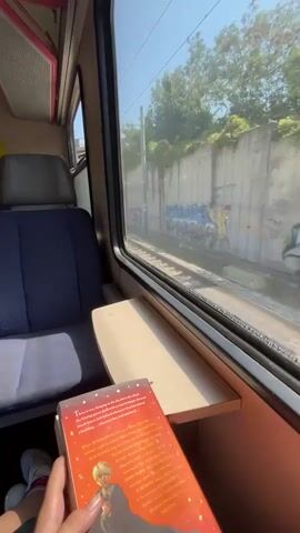 The video begins with a close-up shot of a person's hands holding a book with an orange cover. The camera then pans to the left, revealing the interior of a train carriage. A blue upholstered seat is visible to the left of the frame, and a small wooden table is positioned in front of it. Through the train window, a landscape is visible, consisting of a railway track, a concrete wall with graffiti, and trees. The train appears to be in motion, as indicated by the blurring of the landscape outside. The sky is clear and blue, suggesting a sunny day. The video continues to show the changing scenery outside the window as the train moves forward. The book remains in the foreground throughout the clip.
