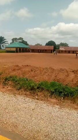 This media file captures a slow, panning view of a dirt road with a gravel shoulder, likely in a rural or semi-rural setting in Madakiya, Nigeria. The scene is dominated by earthy tones of red soil and light-colored gravel.

To the left of the road, a series of buildings are visible. The first appears to be a low-rise structure with a green roof, possibly a community building or school. Further along, there are more buildings constructed from brick or concrete blocks, some appearing unfinished. A prominent palm tree with large fronds stands near one of these structures, indicating a tropical or subtropical climate.

Scattered vegetation, including grass and trees, borders the road and the buildings, suggesting it is daytime with fair weather. The sky is visible in the background, with patches of white clouds against a light blue.

There are no prominent objects on the road itself, though a piece of what appears to be light-colored plastic or trash is visible on the right shoulder of the road. A few indistinct figures of people can be seen in the distance near the buildings, suggesting the area is inhabited and potentially a place of activity, though no specific event or action is clearly depicted. The overall impression is one of a quiet, everyday scene in a developing area. No text is visible in the media file.