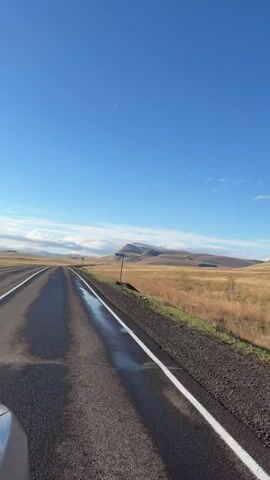 The video depicts a view from a car driving on a straight, two-lane road under a bright blue sky with scattered white clouds. The road surface appears to be wet.  On either side of the road is a field of dry, golden-brown grass. In the distance is a mountain with some snow at its peak.  A road sign indicating "no passing" is visible on the right side of the road. The edge of the car's hood is visible in the lower left corner. The overall impression is of a remote, rural landscape.