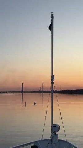 The video shows the bow of a boat sailing on a calm body of water at dawn. A flagpole with a black flag is in the foreground, with a light at its top. In the distance, a bridge with multiple towers spans the water. The sky is a soft gradient of pink and orange, with clear blue above, indicating the sun is just rising. The water reflects the sky and the bridge, creating a serene atmosphere. A single buoy is visible in the water. The boat is moving forward, as evidenced by the slight motion blur and the perspective.