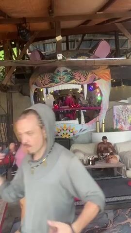 The video shows an indoor space with a wooden roof, where a DJ booth is integrated into a large, colorful tiger head sculpture. The DJs are visible inside the tiger's mouth, surrounded by lights. Below the structure are people relaxing on beanbag chairs and a sofa. One woman is reaching for something on a low table. A man is sitting on the sofa looking at his phone. The space is decorated with rugs and colorful artwork. A hammock chair hangs to the side.