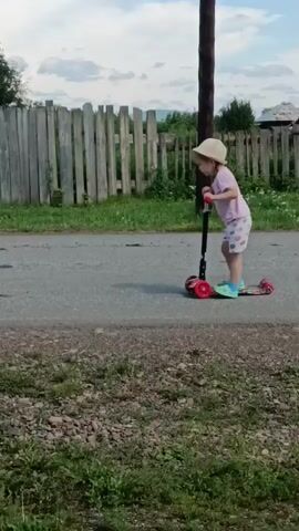 A young child wearing a hat, pink shirt, and patterned shorts rides a colorful scooter down a paved road. A wooden fence runs along the side of the road, behind which are trees and houses. A utility pole stands tall next to the fence. In the background, a two-story house with a red roof and light-colored siding is visible, with a black and white cow standing near a red fence and a quad bike.
