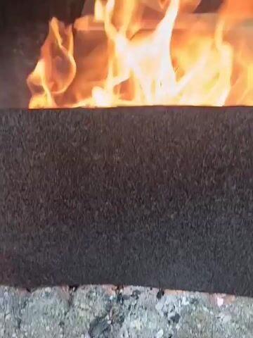 The video shows a close-up of a fire in a hearth or fireplace. There are flames actively burning. A piece of wood is visible amidst the fire, likely being consumed by the flames. The hearth itself appears to be made of a dark, textured material. The overall environment is dark, highlighting the intense orange and yellow of the flames.