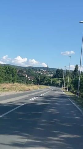 The video presents a forward-facing perspective from a vehicle moving along a paved two-lane road on a clear, sunny day. The road has a double white line marking its center and shows patches of asphalt. Streetlights are visible along the right side of the road, with trees and green foliage bordering both sides. In the mid-ground and background, a hilly landscape covered with houses and trees is visible, with a distinctive tall tower prominently positioned on a distant hilltop. Two cars traveling in the opposite direction pass the camera's vehicle. Long shadows are cast across the road from the right side, indicating bright sunlight.