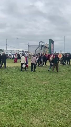 A group of people are gathered in an open grassy field under a cloudy sky. In the background, there is a stage with a large screen and scaffolding. Some people are wearing traditional clothing with red and black designs. Others are dressed in casual modern attire. There appears to be a fair or festival taking place, with a yellow fence visible on the right side of the frame. Several individuals are walking around the field, some are standing in groups, and others are moving towards or away from the stage area. The overall atmosphere seems to be one of a public gathering or event.