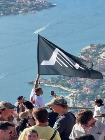 A group of people are gathered on an elevated viewpoint overlooking a bay with several boats. The focal point is a man holding a black flag with a white mountain-like design and a star on the right side. The flag is partially unfurled and angled towards the right. The man is wearing a white t-shirt with a red graphic. Several other individuals are visible in the foreground, some holding phones, appearing to take pictures or record videos. The environment is sunny, with a clear blue sky and a vast expanse of water. The background features a coastal town with red-tiled roofs nestled along the shoreline, and lush green hills in the distance. The water is calm with white wakes trailing behind the moving boats.