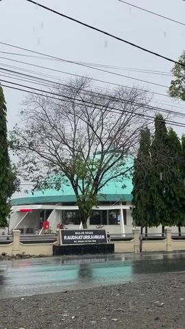 The video shows a large deciduous tree in front of a building with a teal roof. The building has a sign that reads "MASJID BESAR RAUDHATURRAHMAN." It is raining, and the ground is wet and reflects the surrounding objects. There are power lines overhead. The tree is bare of leaves except for some green foliage at the bottom. Two evergreen trees are visible on the right side of the frame. The overall scene appears to be a street view of a mosque on a rainy day.