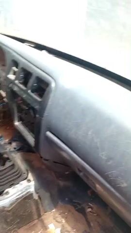 The video shows the interior of a car, likely a truck or utility vehicle, that appears to be in disrepair or undergoing some form of dismantling or modification. The dashboard is visible, showing a cracked windshield and a heavily scratched plastic surface. To the left, a steering wheel is present, though the steering column seems to have been tampered with, with wires exposed. The center console reveals a manual gear shifter, suggesting a manual transmission. The floor of the vehicle is cluttered with debris, including what appears to be torn fabric and possibly insulation or old carpeting. The seats are visibly worn and stained, with one seat showing dark material affixed to it. The overall environment suggests a neglected or functional workspace rather than a typical passenger car interior. The background outside the car, visible through the windshield and door, appears to be an outdoor setting with buildings and laundry hanging.
