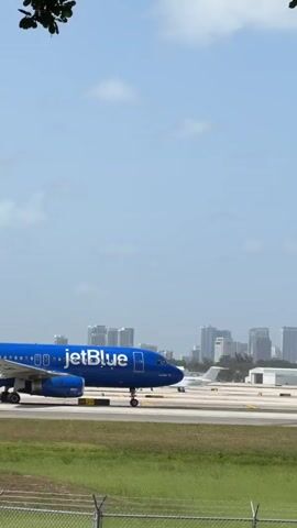A JetBlue airplane with a blue and white livery is on a runway. The plane is a narrow-body jet, possibly an Airbus A220 or A320 series, indicated by its engine placement and wing design. The JetBlue logo is visible on the fuselage. The airplane is facing to the right of the frame and appears to be taxiing or preparing for takeoff or landing. The background shows a cityscape with buildings and a clear sky with some clouds. In the foreground, there is a grassy area and a metal fence. The scene is framed by the branches and trunk of a tree, with foliage obscuring parts of the view. The lighting suggests it is daytime with sunlight.