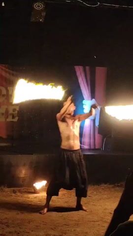 A shirtless man with dark pants is performing a fire dance in front of a dark background. He is holding two flaming objects, one in each hand, and swinging them in a circular motion. There are flames on the ground to his left and right. Behind him, there is a stage with red curtains and a cartoon hand pulling them open. A projection screen is visible above the curtains. The lighting is dark, with the flames providing most of the illumination. The ground appears to be dirt or sand. There are also blurry figures of people in the foreground, suggesting an audience.