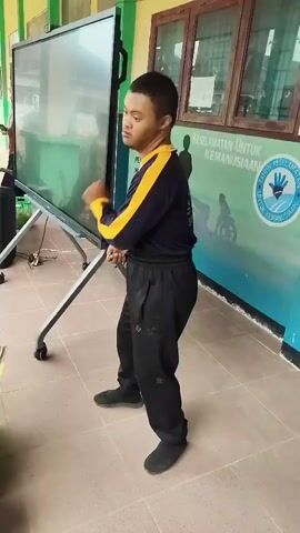 A young man with Down syndrome stands facing a large interactive screen, his hands in a boxer's stance. He is wearing a navy blue long-sleeved shirt with yellow accents on the collar and sleeves, and black sweatpants. He is barefoot. The screen is on a silver wheeled stand and is angled slightly. In the background, a wall features a green and blue mural with text and an image of a hand and a person in a wheelchair. Wooden window frames are visible above the mural. The floor is tiled. The man appears to be in motion, possibly mimicking a dance or exercise.