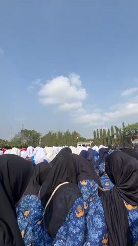 The video shows a large group of people, many wearing blue shirts with a gold emblem and black headscarves, bowing their heads in unison. The scene appears to be an outdoor gathering, possibly a ceremony or prayer service. The sky above is blue with scattered white clouds. In the background, there are trees, a white tent or structure, and an Indonesian flag. The people are positioned in rows, facing the same direction, and their posture suggests a solemn or respectful act. The focus is on the backs of the heads of the individuals in the foreground, obscuring their faces.