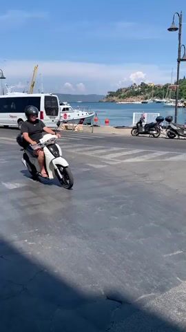 A person wearing a black t-shirt, shorts, flip-flops, and a black helmet rides a white scooter with a black top case on a paved road. The scooter has a license plate that reads "X6T 171P". The rider is looking forward and appears to be moving. The setting is a coastal town with buildings and a body of water in the background. Several other scooters are parked in the background.

The scene is set in Porto Santo Stefano, Italy, during the daytime under a clear blue sky with bright sunlight, suggesting warm weather. The architecture of the buildings, with their pastel colors and balconies, is typical of Italian coastal towns. In the distance, yachts are visible docked in a harbor, indicating a port or marina.

There are several signs visible: a blue "P" for parking, a circular red sign with a horizontal white bar, and a blue directional sign with an arrow pointing right and text that reads "Aurelia 14 SS 1". There is also a sign with a red circle and "30" inside, indicating a speed limit of 30 km/h. A shop sign reads "TRATTORIA".

The overall mood appears to be relaxed and leisurely, consistent with a vacation or tourist destination. The paved road shows some cracking and wear, and there are streetlights and a trash can visible along the roadside. The presence of other vehicles and pedestrians suggests a lively town environment.