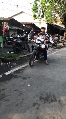The video shows a street scene in Ngaban, East Java, likely in the late afternoon given the warm, long shadows cast by the trees. The weather appears to be clear and sunny. The foreground features a man in a striped shirt and dark pants, riding a motorcycle. He is carrying a white container with a red lid in his left hand. To his right, parked along the side of the road, are several other motorcycles, suggesting a busy area, possibly near shops or businesses.

In the background, there are buildings with open fronts, resembling shops or stalls. One building has a white sign with some writing, though it's difficult to read clearly. Another sign above one of the stalls has text in what appears to be Indonesian. A few people are visible within the stalls, some appearing to be attending to customers or working. Trees line the street, providing some shade. The overall atmosphere seems to be one of everyday activity in a commercial street setting. The emotions conveyed are neutral, focusing on the depiction of daily life.