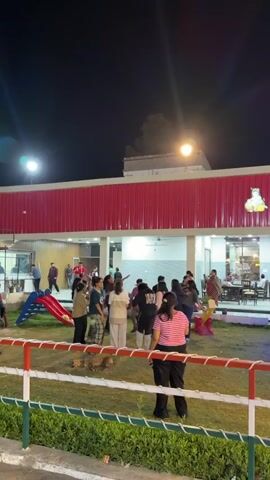 This media file captures a lively outdoor gathering at night, likely a party or celebration, in Gannaur, India. The scene is set in a spacious area with a building featuring a prominent red corrugated metal facade. In front of the building, a well-lit grassy area is occupied by a group of people, predominantly young adults, engaged in various activities.

The central focus appears to be a group of individuals dancing and interacting in a circle, suggesting a joyful and festive atmosphere. Their body language and postures convey a sense of energy and enjoyment. Scattered around the area are other people socializing and observing the activities. Visible objects include a children's slide with a red ramp, a white fence with red and green accents, and some potted plants.

The time of day is clearly night, indicated by the dark sky and the artificial lighting from overhead lamps and string lights. There are no visible cues about the weather, but the presence of an outdoor event suggests it is pleasant. The building architecture, with its red metal siding and open-fronted structure, offers some location cues that might be recognizable to someone familiar with the area.

Although no specific text is clearly legible within the main activity area, there are some blurred signs and logos on the building in the background that are not decipherable. The overall mood is one of cheerful festivity and social engagement.