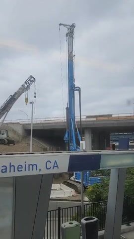 The video shows a construction scene in Anaheim, CA. The foreground features a modern, grey bus stop shelter with a blue and white sign that reads "Anaheim, CA" and an arrow pointing left with "Orange" below it. Two trash receptacles are visible near the shelter.

In the background, a large blue drilling rig stands tall, with a long drill bit suspended from its boom. To the left of the drilling rig, a large white crane is positioned on a mound of earth. The crane has its boom extended upwards, with a hook hanging from it. In the distance, a bridge or overpass under construction is visible, with concrete pillars and structural elements. The ground around the construction area appears to be excavated earth and some form of temporary barrier. Lush green trees can be seen on the right side of the frame. The sky is overcast, suggesting a cloudy day. The overall scene depicts ongoing infrastructure development, likely related to transportation. There are no people visible in the video. The time of day is not clearly indicated, but the lighting suggests it is daytime.