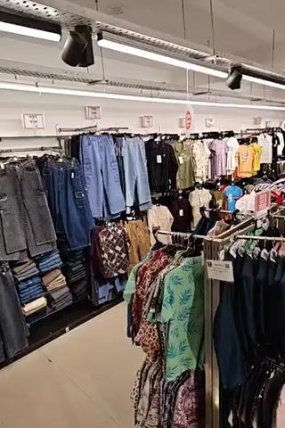 The video shows the interior of a clothing store, with racks of apparel lining both sides of a central aisle. The store appears to be well-lit with fluorescent lighting. On the left side of the aisle, there are racks of jeans, pants, and patterned shirts. On the right side, there are more racks of shirts, dresses, and other clothing items. Several mannequins are visible, some displaying children's clothing. The store seems to be arranged in sections, with different types of clothing displayed together. There are signs visible, some of which appear to be promotional. The overall impression is of a busy retail environment.