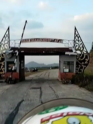 The video opens with a view of an entrance gate for "AGRO WISATA MARGO LAWU". Beyond the gate, a road curves uphill. The camera then pans to a scenic view of a sunset over rolling hills. The sky is filled with soft clouds tinged with orange and yellow hues. The foreground shows dry grass and some scattered bushes. The camera then briefly shows the interior of a dark room with a window reflecting the sunset colors. Finally, the camera returns to the panoramic view of the sunset over the hills and vegetation.