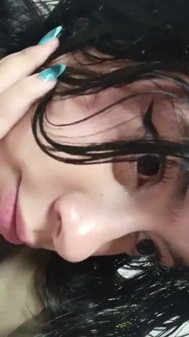 The video presents a close-up shot of a person's face. The individual has long, dark, and somewhat disheveled hair, which appears to be damp or wet. They possess a fair complexion, dark eyes, and a slight, gentle smile. A hand with long, bright blue, manicured fingernails is visible in the frame, occasionally touching the hair near the side of the face. The person looks directly at the camera. The setting appears to be indoors, under artificial lighting. No specific location cues are discernible within the frame. No text is visible.