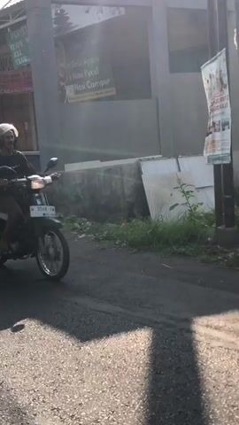 The video shows a side view of a person riding a motorcycle. The motorcycle is carrying a green woven basket on the back, filled with white bags and some items that appear to be food. Behind the basket, there is a large white plastic bag and a black plastic bag. The rider is wearing a helmet. In the background, there is a gray building, a tall pole with wires, a tree, and a green field. Later in the video, another motorcycle carrying two people, a male and a female, passes by. The female is wearing a pink sweater and blue jeans, and the male is wearing a black sweater. The video appears to be taken during the day with bright sunlight casting shadows.