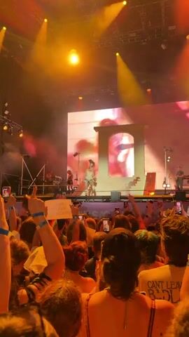The video shows a concert with a performer on stage, illuminated by bright orange lights and spotlights. The performer is standing in the center of the stage, wearing light-colored clothing. Behind the performer is a large screen displaying a close-up of a person's face, partially obscured by a decorative archway. In front of the stage, a large crowd of people is visible, many holding up their phones to record the performance. The audience is bathed in the same orange light as the stage, creating a warm and vibrant atmosphere. There are some smoke effects on stage, adding to the dramatic visual. The overall impression is a dynamic and energetic live music event.