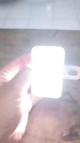The video shows a close-up view of a hand holding a rectangular, white device with a white loop attached to its right side. The device is emitting a bright, white light. The hand, with visible fingers and fingernails, is partially obscuring the light from the left side. The person's fingertip is pressing down on the surface of the device. The background appears to be a wooden table with a tiled surface in the upper left corner. The lighting in the scene suggests it is indoors.