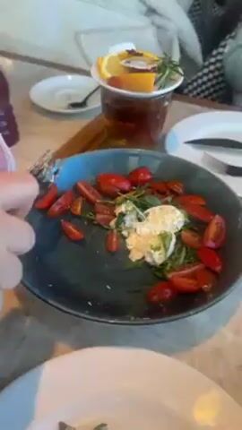 The video shows a meal at what appears to be a restaurant.  A plate with cherry tomatoes, cheese (possibly burrata), and arugula salad is visible.  A fork is used to take a bite. A basket of bread is also visible.  There's a close-up of someone’s hand reaching for bread.  Next, a white plate appears with a slice of bread topped with cheese, tomatoes, and arugula. A napkin and knife are on the table next to the plate. A beer glass and bottles are visible in the background.