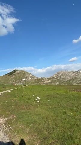 The video shows a green, grassy field with rocky hills in the background under a blue sky with white clouds. As the camera pans, a white car is seen driving along a road on the edge of a hill overlooking a mountainous landscape. The sun shines brightly behind a large cloud.