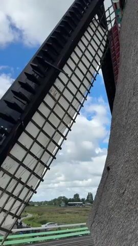 A video captures a close-up view of a windmill blade. The blade is made of wood and is painted red on one side with black latticework. The blade is moving, suggesting the windmill is operational. In the background, a picturesque Dutch landscape unfolds with other windmills, houses, and a canal under a blue sky with white clouds. People are visible walking along a path near the canal. The camera perspective is from within the windmill structure, looking outwards. The overall scene evokes a sense of rural Dutch scenery.