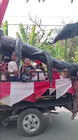 Children and adults are riding in the back of a black pickup truck. The truck bed is decorated with red and white fabric. A large, black, scorpion-like float is on the truck, with children and adults interacting with it. A woman in a hijab is standing next to the truck and appears to be adjusting the float. The background shows banana trees and some buildings. The overall scene appears to be a parade or festive event.