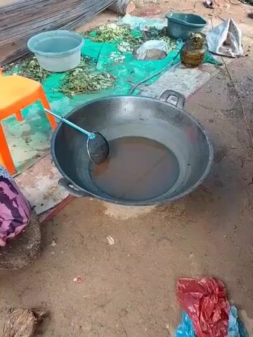 A video captures an outdoor scene with a large metal wok filled with oil, resting on the ground. A metal slotted spoon is partially submerged in the oil. To the left, a portion of a person's leg, clad in patterned fabric, is visible, along with a small stool. In the background, a pile of green foliage is spread out on a blue tarp, and various containers and objects, including rebar and plastic bags, are scattered around. The ground appears to be dirt and some concrete.