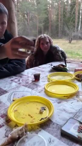 The video shows three people seated at a table laden with food and drinks under a wooden shelter. The background shows a forest setting. The camera focuses on a woman with curly hair seated directly across the table, who quickly turns away and stands up. Then the camera shifts to a man who raises a small glass toward the camera. The table displays several yellow plates, bread, a container of red liquid, a box of cookies, and other items. The man is wearing a dark blue jacket over a red shirt. The atmosphere appears informal and relaxed, potentially a picnic or casual gathering in the woods.