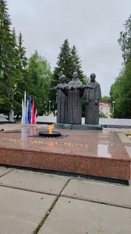 The video displays a close-up view of a memorial with three tall, dark statues of individuals wearing robes and facing forward. Their arms are outstretched, and they appear to be holding or reaching towards something unseen. In the foreground, an eternal flame burns in a bowl, surrounded by a red granite base. Several flags, predominantly white, blue, and red, are visible to the left of the flame. The memorial is set in an outdoor environment with green trees in the background and a cloudy sky overhead. The ground is paved with square concrete tiles. The overall scene suggests a solemn and commemorative setting.