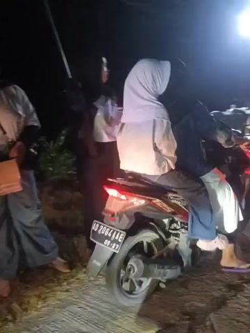 The video captures a nighttime scene with several people gathered around motorcycles on a road. A prominent motorcycle in the foreground has its brake light illuminated, suggesting it is either stopped or braking. Two individuals are seated on this motorcycle, while another person stands nearby to the left. In the background, more people are visible, some standing and others on motorcycles. The environment appears to be outdoors, with a faint light source visible in the upper right portion of the frame, casting shadows on the road and the surrounding area. The road surface is visible and appears to be paved. The overall impression is of a group of people congregated on a road at night, with motorcycles being a central element.