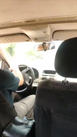 The video is from inside a car. The driver's hands are on the steering wheel. The car is moving on a road surrounded by trees. The camera is angled to show both the interior of the car and the outside scenery. The perspective suggests the camera is positioned in the back seat or passenger seat, looking forward.