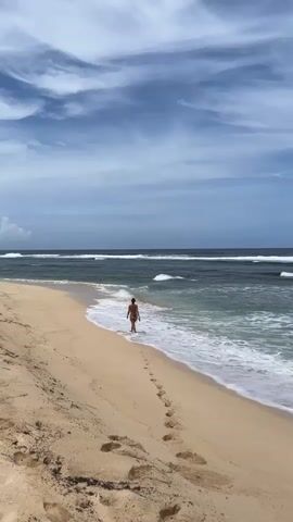 A woman in a swimsuit walks along a sandy beach toward the ocean. The sand is light-colored and shows numerous footprints. The water is teal and blue, with small waves breaking along the shoreline.  The sky is blue with streaky white clouds. The beach appears relatively deserted.
