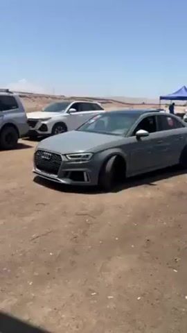 The video shows a gray Audi A3 sedan parked on a dirt surface. The car has black rims and a black roof, and there's a number "366" displayed on the rear side window. The driver is visible inside, wearing a hat and looking towards the camera. Several other vehicles are parked nearby in the background, including a white SUV and a dark SUV. The setting appears to be an outdoor event, possibly a racing event. The weather is sunny with a clear blue sky.