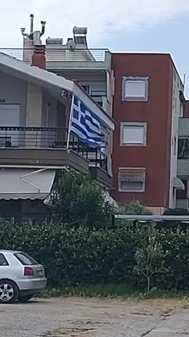 The video displays an exterior view of a residential building with a Greek flag displayed on a balcony. The building features multiple stories, balconies, and an air conditioning unit mounted on the exterior wall. A silver car is parked in front of a hedge. The scene appears to be in a residential neighborhood with trees and a fence.