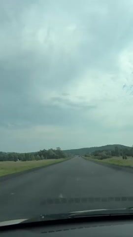 This video shows a first-person perspective from inside a vehicle traveling on a paved road. The sky is overcast with clouds, and the road stretches into the distance, surrounded by green fields and trees. The dashboard of the vehicle is visible at the bottom of the frame. There are no other vehicles or people clearly visible on the road. The environment appears to be rural.