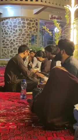 A group of five men are sitting on a red rug on the ground, possibly outdoors. They appear to be in a semi-circle, facing each other, and engaged in conversation. Two of the men in the foreground are wearing brown traditional clothing. One of them is holding a phone. Another man is wearing a grey shirt and has a beard. The background shows a building with stone walls and lights. There is a bottle of water on the rug.