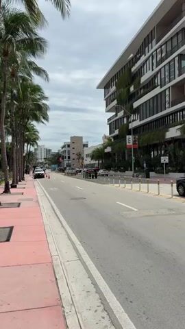 The video opens with a series of street-level views, showcasing a lively urban setting, likely Miami Beach. Palm trees line the red sidewalks, flanking a road with several vehicles, including SUVs and sedans. Modern, multi-story buildings with glass facades and some greenery on balconies dominate the background. The sky is overcast but bright, indicating daytime. Yellow bollards separate parked cars from the main road in some sections. One shot clearly displays a red circular "No U-turn" sign.

The scene then shifts to a selfie video featuring a young man, possibly in his late teens or early twenties. He is wearing a white long-sleeved shirt and a camouflage baseball cap adorned with a distinct symbol on the front. With a wide, excited smile, he winks one eye and makes a "rock on" hand gesture, extending his index and pinky fingers. He enthusiastically announces, "We're here in Miami, let's get it!" His energetic expression conveys a sense of excitement and enjoyment.