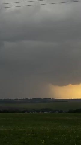 The video shows a wide shot of a green field in the foreground, a distant horizon line of trees and possibly a few buildings, and a dramatic sky. The sky is predominantly filled with dark grey storm clouds, but a pocket of golden sunlight breaks through on the right side, illuminating part of the cloud cover and possibly the distant landscape. It is raining in the distance. Powerlines are visible at the top of the frame. Music is playing.