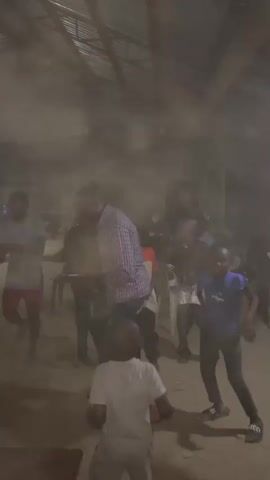 This media file captures a lively gathering of people, likely a celebration or social event, taking place indoors. The scene is somewhat dimly lit, with a corrugated metal roof suggesting a possibly rustic or unfinished building. Several individuals, including adults and children, are actively engaged in dancing and interacting. Some are captured mid-motion, their bodies conveying a sense of energy and enjoyment. Objects such as tables and chairs are visible, indicating a communal space.

The atmosphere appears to be festive, with participants expressing joy and enthusiasm through their movements. The presence of children suggests a family-friendly or community-oriented event. The overall ambiance is one of shared celebration and active participation.

While the exact time of day is not explicitly stated, the indoor setting and lighting might imply an evening or late afternoon event. There are no clear indications of weather conditions, though the indoor setting suggests protection from the elements. The mention of Nigeria as a location context provides a geographical anchor to the scene. There is no visible text in the provided frames.