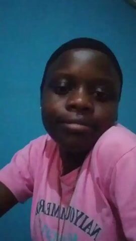 A person with dark skin is facing the camera against a blue wall. They are wearing a pink t-shirt and a black hair covering. A thin silver chain is visible around their neck. The person's facial expression is mostly neutral with occasional subtle smiles and puffed cheeks. The lighting in the room appears consistent, illuminating the subject evenly.