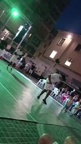 Two men in white shirts and shorts are on a basketball court. One man is in the foreground and is bent over, looking down. The other man is in the background and is facing away from the camera. There are spectators sitting in chairs on the right side of the court. There are buildings in the background and lights are shining on the court.