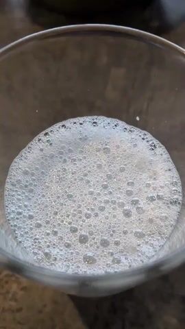 A close-up shot shows the surface of a clear liquid in a glass or jar. The liquid is filled with numerous small, white bubbles, creating a foamy texture. The bubbles are of varying sizes, with many appearing to be smaller than a pea. The liquid itself is clear and transparent, with the bubbles obscuring the bottom of the container. The background is out of focus but appears to be a countertop or table surface. The lighting suggests an indoor environment.