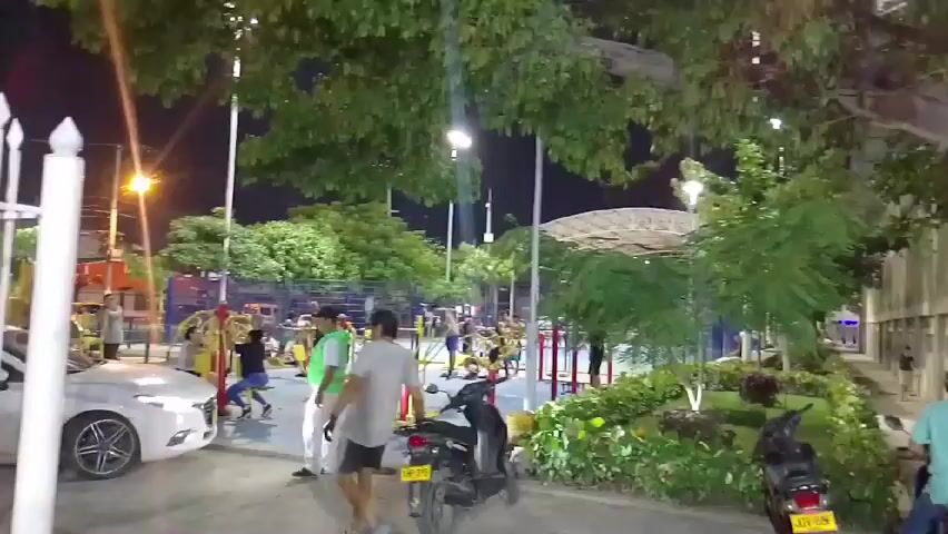The scene takes place at night in a well-lit outdoor park area with exercise equipment. Several people are using the equipment for exercise. Two scooters are parked on the ground near a small garden area. A white car is parked on the left side of the frame. Streetlights illuminate the scene. A man wearing a green vest walks towards the exercise equipment. A building with an arched roof is visible in the background.